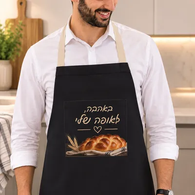 גבר חובש כיפה לובש סינר כותנה שחור עם הדפס חלה ועיצוב "באהבה, לאופה שלי" במטבח מואר